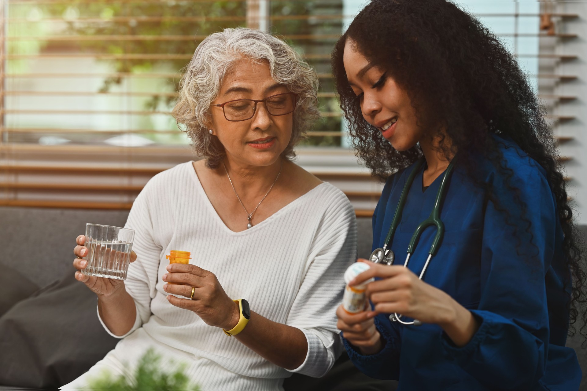 A Young African nurse or caregiver in a scrubs uniform showing a medicine bottle to remind an elderly Asian woman to monitor, Home visit care nursing service for the elderly concept